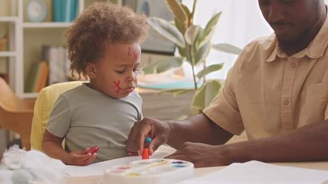 Cute toddler girl with painted face and her loving father drawing together with wax crayons at home