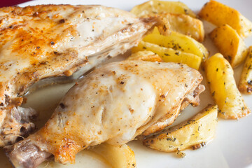 A juicy chicken thigh with breast and baked potatoes with rosemary in the background on a white plate. Traditional food.