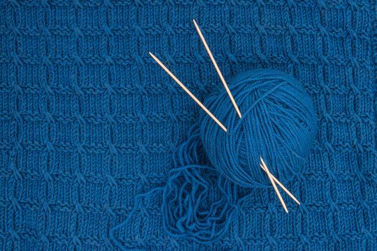 A Ball Of Blue Yarn And Two Knitting Needles On A Background Of Blue Knitwear. View From Above