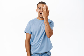 Enthusiastic young man hides one side of face, covers an eye and smiles, stands over white background