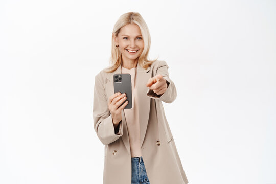 Elegant, beautiful middle aged woman recording video on smartphone, taking photos on mobile phone camera, smiling, standing over white background