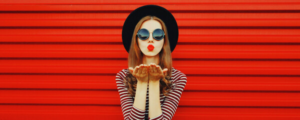 Portrait of stylish young woman blowing her red lips sending air kiss wearing black round hat on...