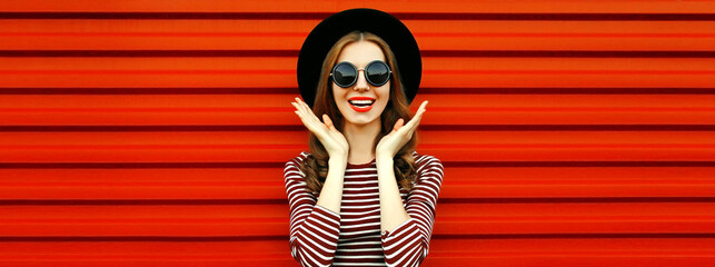 Portrait of happy excited young woman wearing black round hat and sunglasses on red background