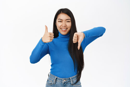 Like or dislike. Smiling asian girl shows thumbs up and thumbs down, makes choice, rate and give feedback, makes decision, stands over white background