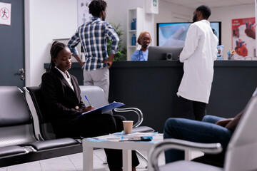 Young adult writing medical report papers and checkup form to receive healthcare insurance support before appointment with physician. Patient sitting in waiting room area at hospital facility lobby.