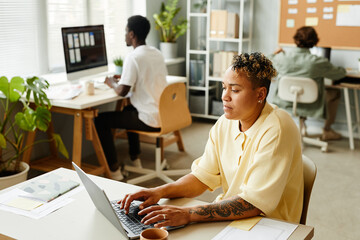 Portrait of tattooed black woman using laptop in office while working with team in background, copy...