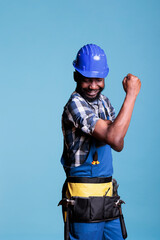 Strong man flexing arm muscles, preparing to work on construction reform with tools. Confident and energetic african american builder pointing biceps and triceps, studio shot against blue background.