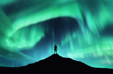 Northern lights and silhouette of standing man on the top of mountain. Aurora borealis and happy guy. Sky with stars and green polar lights. Colorful night landscape with aurora background. Concept
