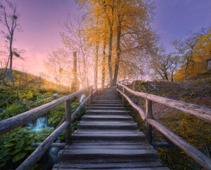 Fototapeta na wymiar Wooden stairs in forest at sunset in autumn. Plitvice Lakes, Croatia. Colorful landscape with path in park, steps, yelllow trees, water lilies, river, pink sky in fall. Trail in woods. Nature