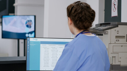 Medical nurse checking exam appointments on computer at hospital reception desk, working on checkup visits with report papers and forms. Doing healthcare insurance work in facility lobby.
