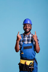 African american construction worker wearing work clothes and hard hat pointing upward with both hands. Happy construction worker posing for camera in studio shot against blue background.
