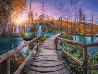Waterfall and wooden path in orange forest in Plitvice Lakes, Croatia at sunset in autumn. Colorful landscape with trail in park, trees, water lilies, river, pink sky in fall. Trail in woods. Nature