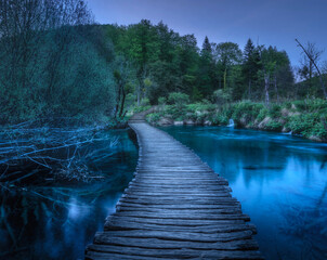 Beautiful wooden path in green forest in Plitvice Lakes, Croatia at dusk in spring. Colorful landscape with trail in blooming park, trees, river, blue sky in summer at night. Trail in woods. Nature