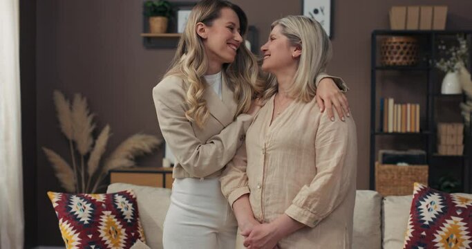 Loving adult daughter hugs middle-aged mother, they look into the camera, happy mature grandmother and granddaughter laugh at each other.