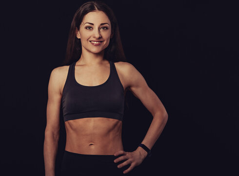 Confident sportswoman in grey sportsbra and shirt, holding hands on waist, fitness trainer standing in power pose, workout in gym isolated on black background