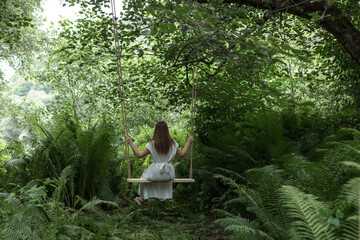 A young girl in a white dress rides on a rope swing in the wood. The concept of outdoor recreation