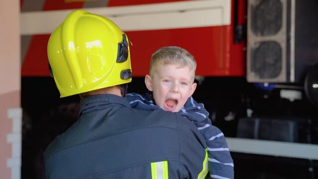 Brave firefighter in uniform holding little funny boy against the background of a fire engine