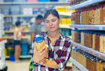 Interested young woman standing near shelves with jars of canned vegetables in supermarket, choosing beans in sauce