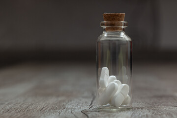 Glass container with white pills on a wooden table. the Container is located to the left of the center picture on a grey background. 