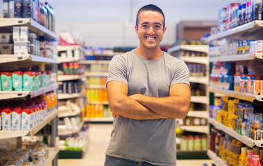 Portrait of positive caucasian man with arms crossed standing in aisle of supermarket, smiling and looking at camera.