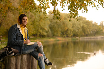 Young creative man with dreadlocks drawing, writing in notebook, sitting on stumps near lake in parkland.