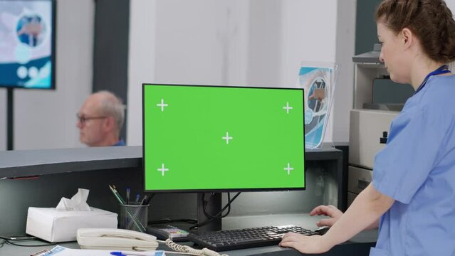 Medical nurse working with greenscreen on computer at hospital reception desk. Using isolated chroma key background with mockup display and blank copyspace template on monitor.