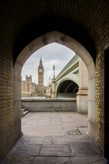 View of Big Ben, Palace of Wesminster and Westminster Bridge with cloudy sky background through an archway under the bridge.