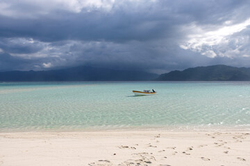 Fototapeta na wymiar A small motorboat moored in the crystal clear turquoise ocean water of a remote sandbar island, rainstorm and dark clouds in distance, tropics of Bougainville, Papua New Guinea