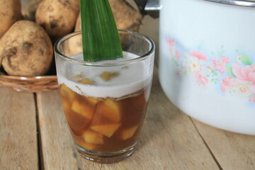 A traditional Indonesian food called jenang ketela in a white glass made of cassava, brown sugar, and tapioca flour with coconut milk topping with pandan leaves as decoration placed on a wooden table

