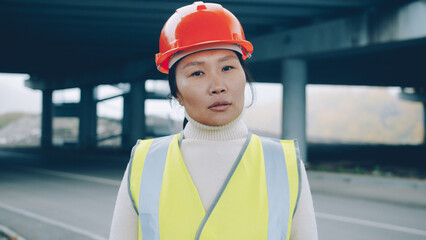 Portrait of serious Asian woman wearing construction uniform stanidng in building zone alone and looking at camera with confident expression