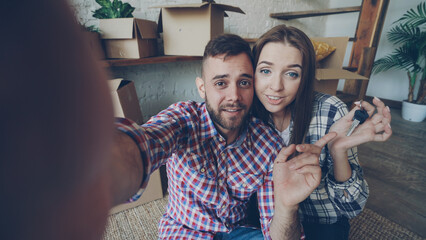 Happy young couple is making video call with smartphone after relocation. They are greeting friends, showing new house keys and many boxes, chatting and smiling.