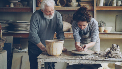 Caring grandfather experienced potter is teaching yong girl how to work with clay on potter's wheel...