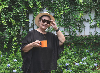 healthy Asian senior woman wearing black blouse, sunglasses and hat, holding orange color cup of coffee, standing  in the garden with purple flowers, , smiling and looking at camera.