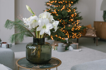 White amaryllis and fir branches in vase on table in stylish modern home interior. Event celebration. Festive holiday decorations. Copy space. Beautiful Christmas tree on background. - obrazy, fototapety, plakaty
