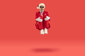 Old cheerful grey-haired man wearing Santa style is sitting levitating in air on red background. He is laughing and looking at camera in dark black sunglasses. Celebrating Christmas, New Year banner.