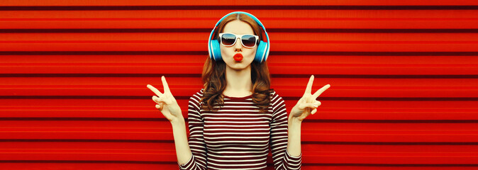Portrait of young woman with headphones listening to music blowing her lips sends air kiss on red...