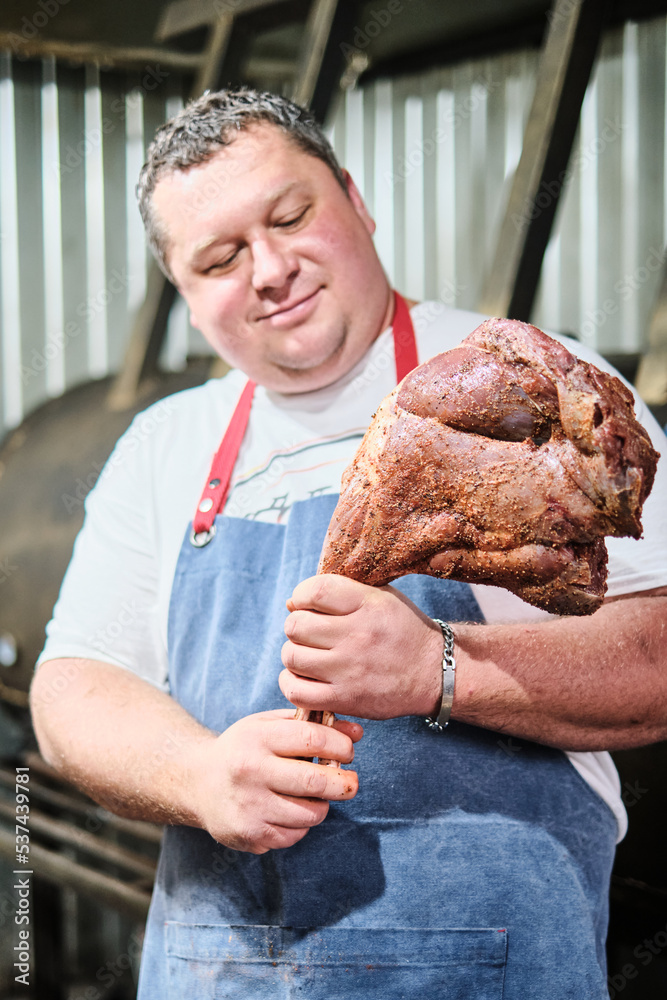 Wall mural the butcher holds a leg of lamb seasoned with marinade in front of a large smoker