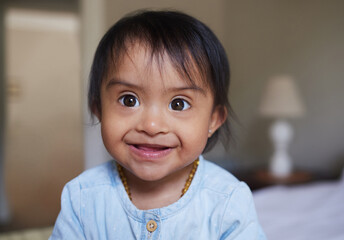 Happy, portrait smile and Down syndrome baby relaxing on a bed in happiness at home. Cheerful little child with genetic disorder or disability smiling in bedroom for cute childhood and development - obrazy, fototapety, plakaty