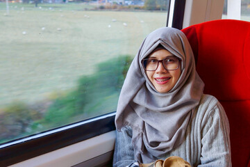 Portrait of young and beautiful Asian Muslim woman smiling and wearing eyeglasses and hijab sitting alone against the window in a moving train. Happy and excited expression. 