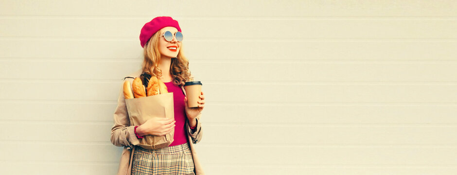Portrait of happy smiling young woman holding grocery shopping paper bag with long white bread baguette and cup of coffee on gray background, blank copy space for advertising text