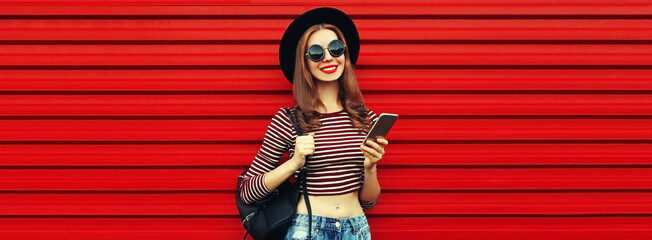 Portrait of stylish young woman with smartphone and backpack wearing black round hat on red background