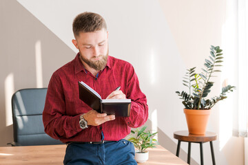 Budget planning. Pensive bearded man writing in a notebook while sitting at the table in a modern office in business clothes, business planning concept