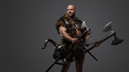 Studio shot of bald viking warrior with fur holding two huge axes.