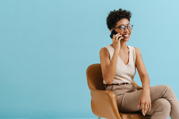 Young curly woman talking on cellphone while sitting in armchair