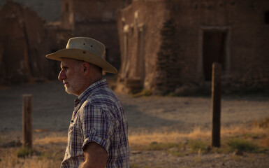 Side view of adult man in cowboy hat and shirt against abandoned building