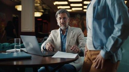 Mature man using laptop, talking with serious businesswoman, showing papers, documents. Business meeting in office restaurant. Signing contract for new economic strategy of company