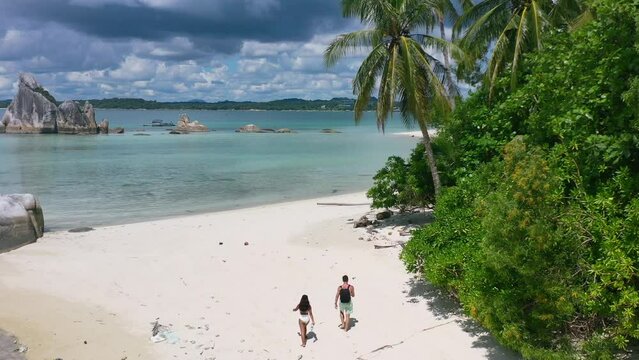 aerial of couple walking on empty white sand beach in belitung indonesia with beautiful tropical ocean
