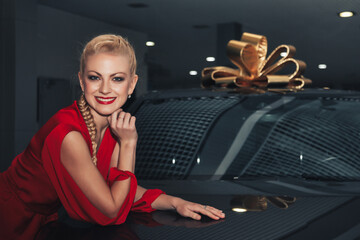 Pretty young woman model smiling and present car, lying on newly bought automobile at dealership. Happy woman in red dress with new auto with gold bow on roof. Car presents concept. Copy text space