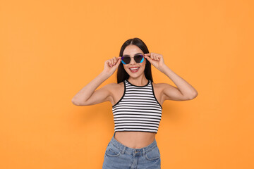 Attractive happy woman touching fashionable sunglasses against orange background