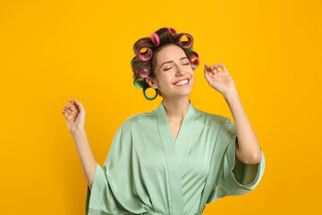 Happy young woman in silk bathrobe with hair curlers dancing on yellow background